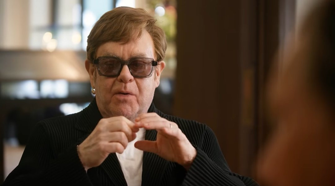 Dressed in a black pinstripe blazer and his signature embellished dark glasses, Elton John animatedly gestures with both hands as he recounts the story of his kneecap surgery to jeweler Theo Fennell in a scene from the documentary short