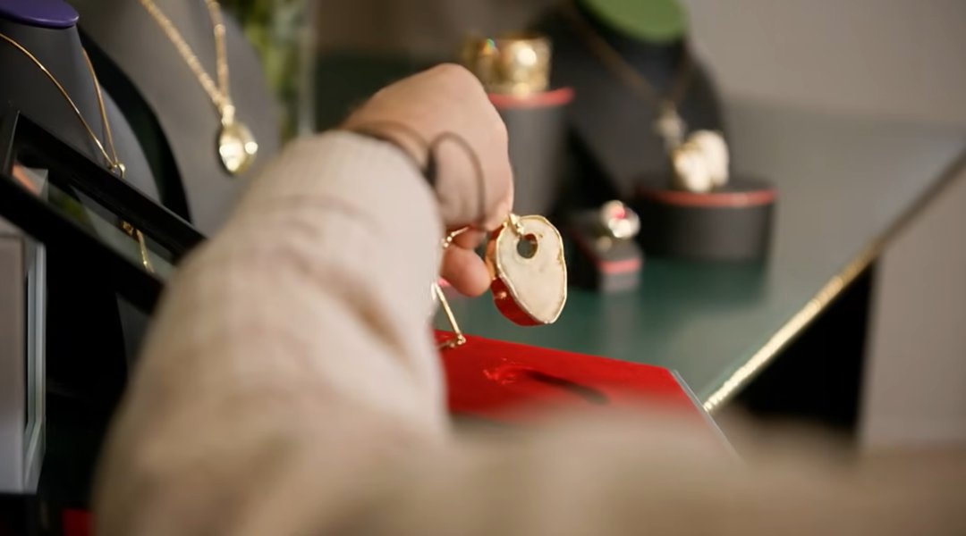 Theo Fennell holds up the finished kneecap necklace — its pale, organic bone pendant edged in gold — over a red jewelry box, with other gold pieces displayed on stands visible in the background of his London workshop. | Source: YouTube/World Gold Council