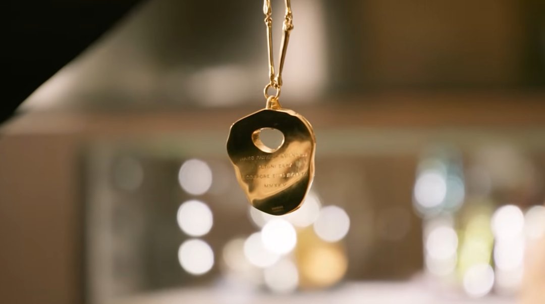 The back of the kneecap necklace, fully gilded in gold, twists on its delicate bone-link chain to reveal the Latin inscription engraved across its uneven surface — catching the light against a soft, bokeh backdrop. | Source: YouTube/World Gold Council