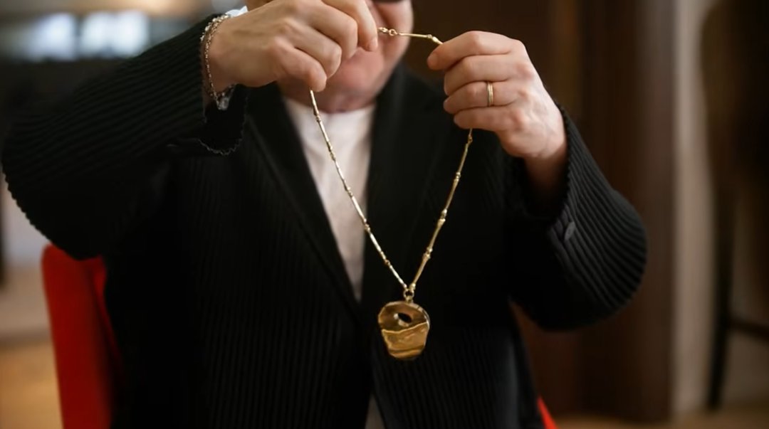 Elton John holds the completed necklace up with both hands, stretching the gold bone-segment chain wide to display the irregular, golden kneecap pendant hanging at its center — the piece's natural hole clearly visible at its core. | Source: YouTube/World Gold Council