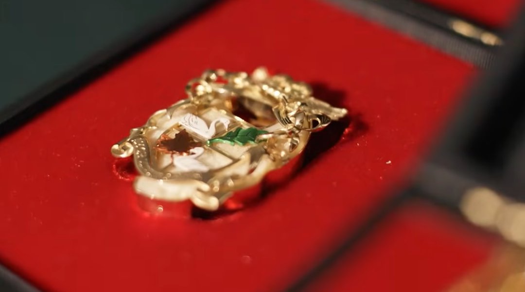 The brooch crafted from Elton John's smaller left kneecap rests on a red velvet jewelry box, its bone fragment encased in an ornate gold setting decorated with intricate detailing in white and green enamel. | Source: YouTube/World Gold Council