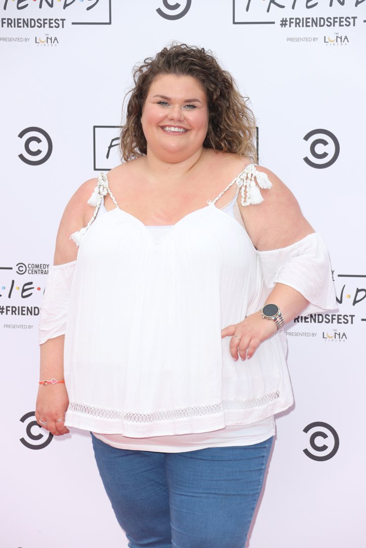 Tapper poses with one hand on her hip against a Friends-branded step-and-repeat backdrop at Comedy Central's FriendsFest on Clapham Common, London, June 2021, smiling warmly into the camera in a flowing white cold-shoulder top and blue jeans.