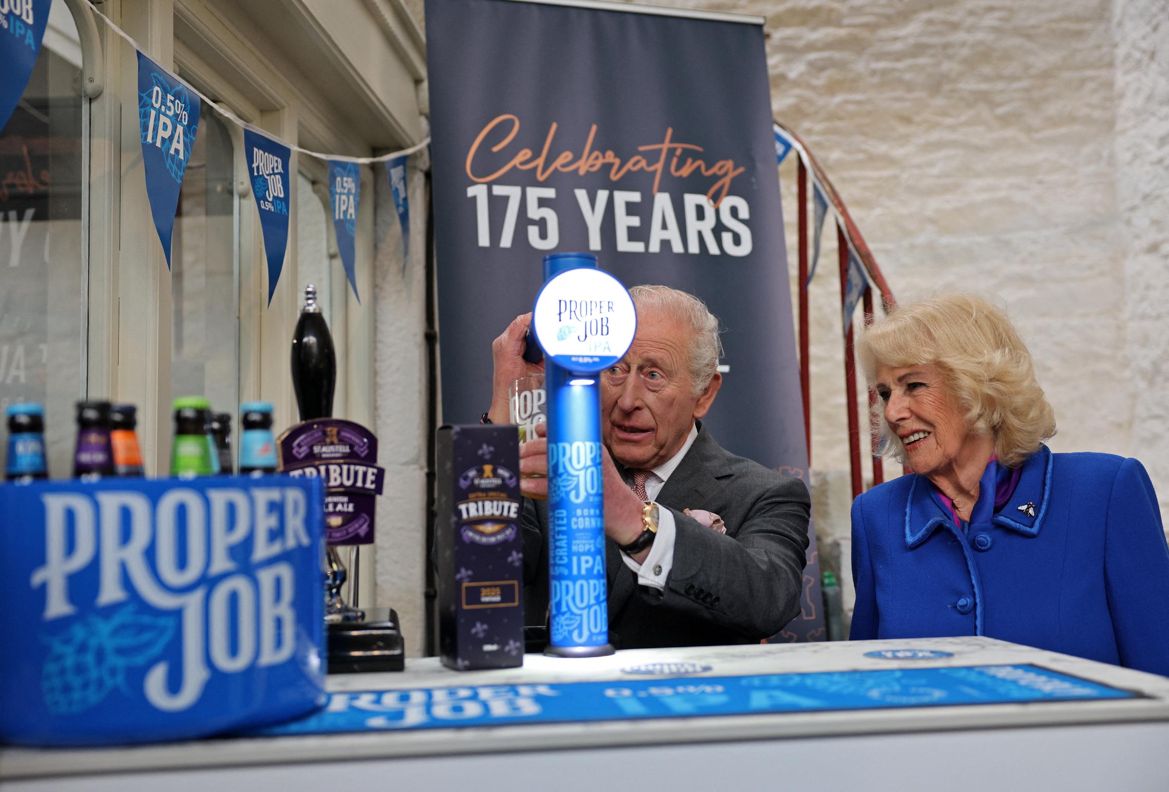 Queen Camilla reacts as King Charles III pulls a pint of St Austell Brewery's Proper Job IPA 0.5% that he pulled, during a visit to the The Market Hall on 24 March 2026 in St Austell, England. | Source: Getty Images
