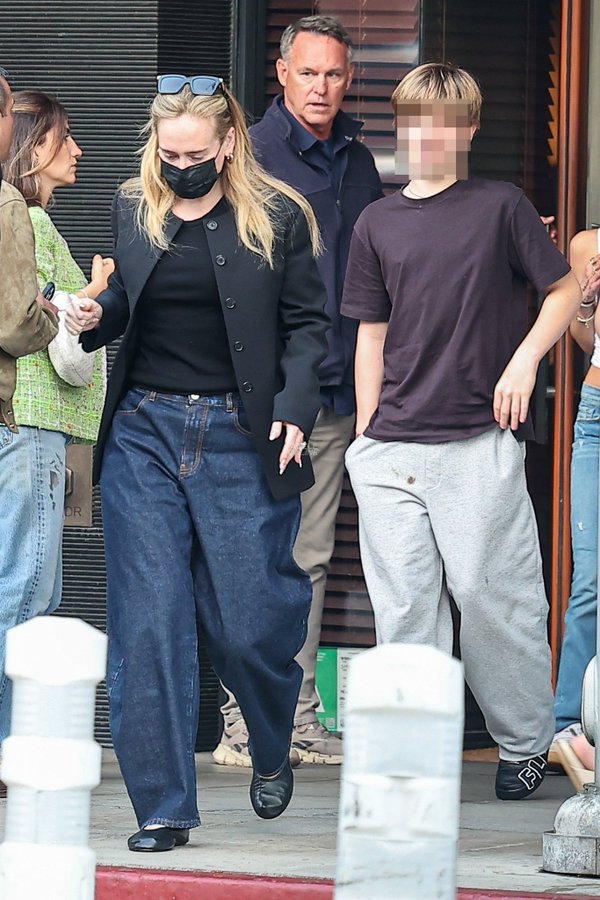 Adele, dressed in wide-leg dark denim, a black jacket, and ballet flats, walks closely alongside her teenage son Angelo &mdash; who sports grey sweatpants and a dark brown tee &mdash; as they navigate their way through a crowd outside South Beverly Grill, with a suited security guard following closely behind. | Source: X/Pop Tingz