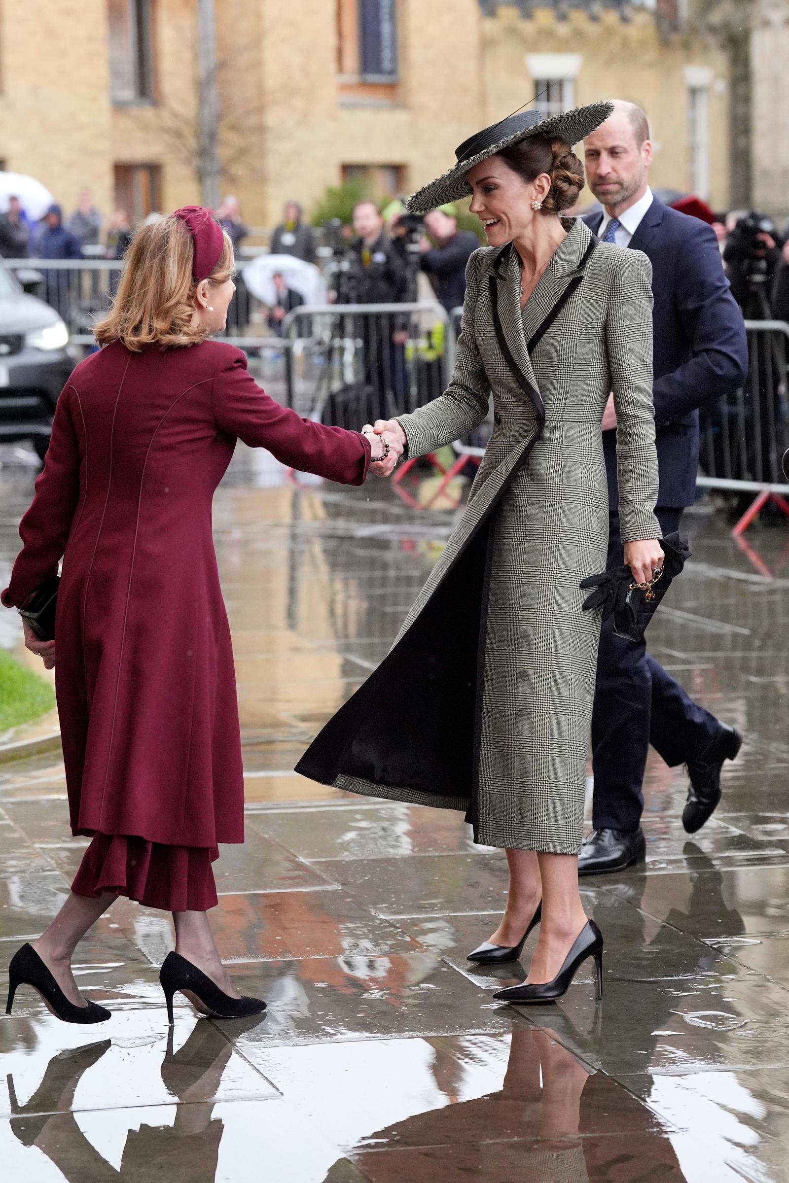 The Princess of Wales, wearing her Suzannah London Prince of Wales check coatdress and Juliette Botterill Millinery boater hat, greets a guest with a warm handshake on the rain-slicked cobblestones outside Canterbury Cathedral on 25 March 2026. Her black Chanel lambskin handbag is visible in her free hand, the gold-tone hardware catching the grey morning light, while her black Ralph Lauren stiletto pumps hold their own admirably against the wet stone beneath her feet.