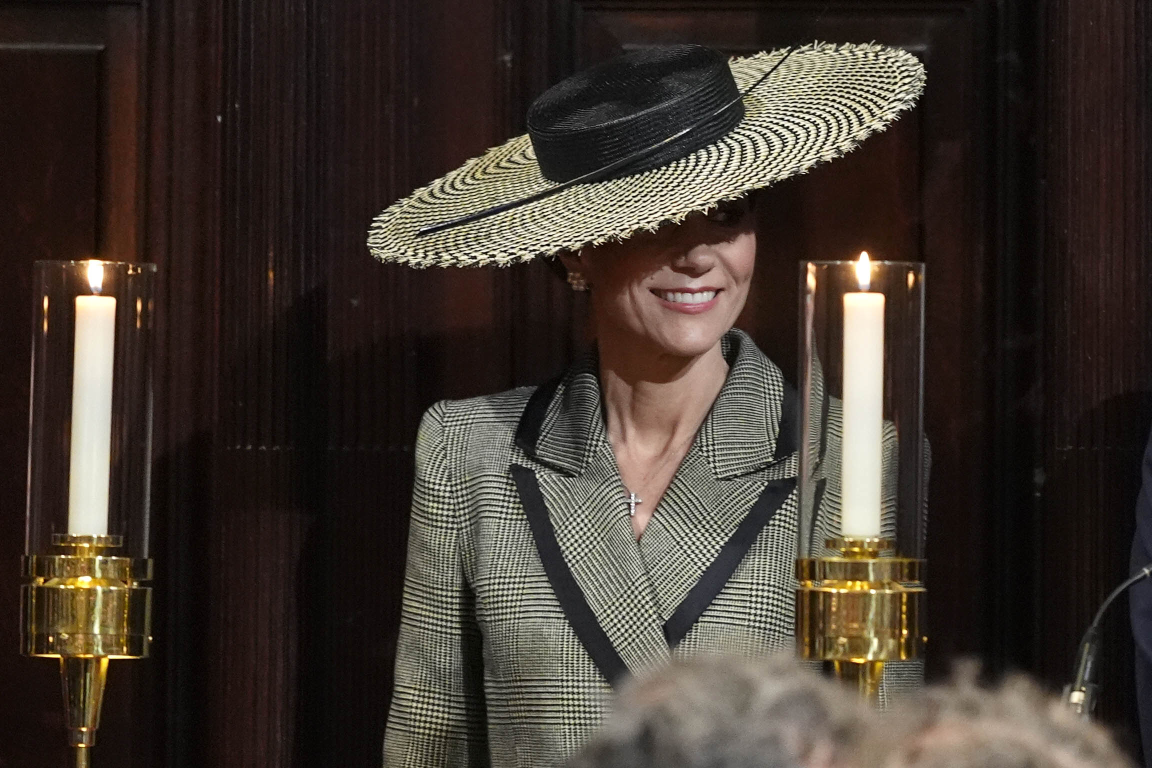 Inside Canterbury Cathedral, the Princess of Wales is flanked by two tall candlesticks, their flames casting a warm glow against the dark wood panelling behind her. The wide houndstooth brim of the Juliette Botterill Millinery boater frames her face almost theatrically in this setting, and the diamond cross necklace is clearly visible at her neckline &mdash; a considered choice for a ceremony marking a historic moment in the Church of England's history.