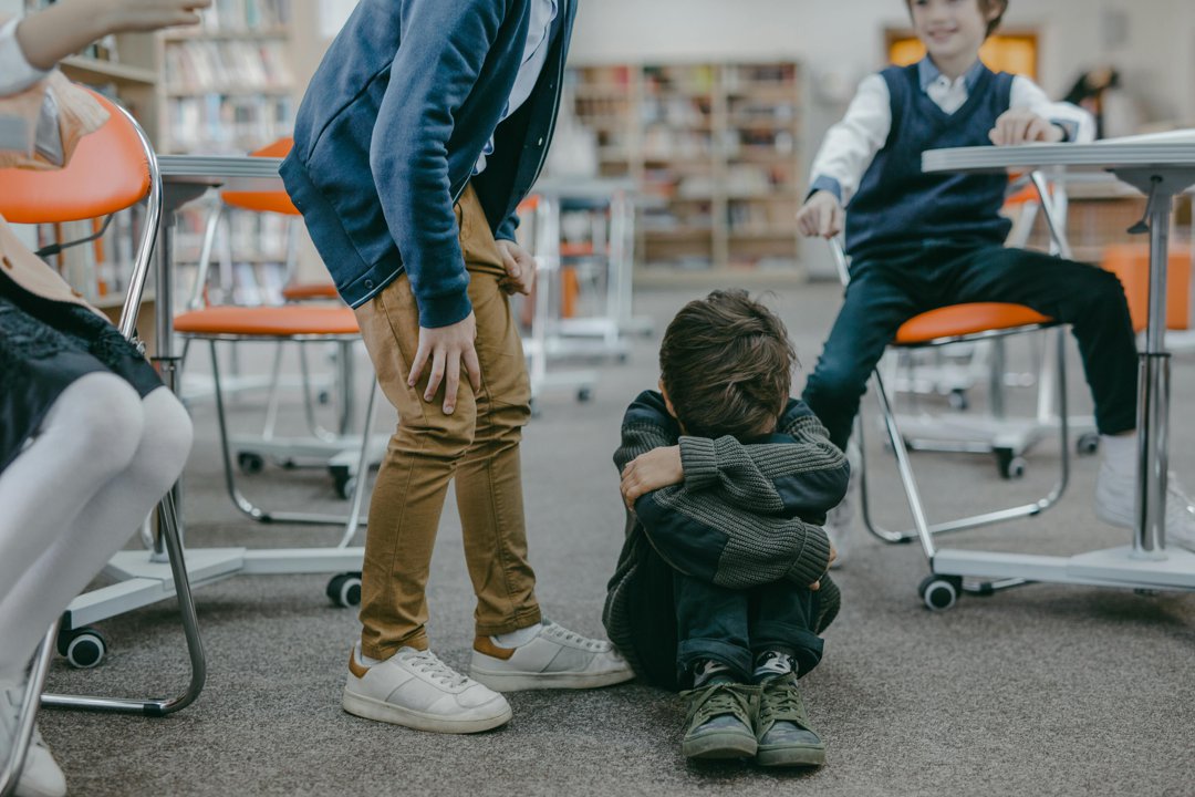 A child sits on the floor of a classroom with their head down as others stand nearby, illustrating the isolation often linked to school bullying. | Source: Pexels