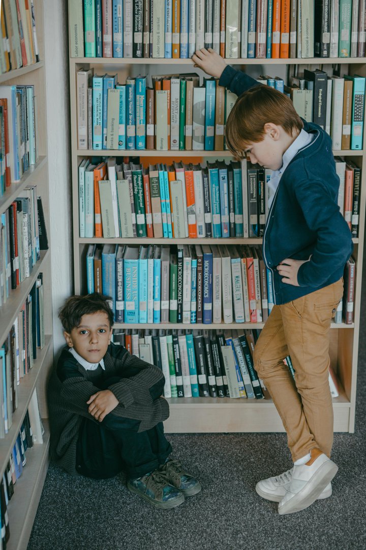 A young boy sits alone beside a bookshelf while another child stands over him, highlighting the imbalance of power often seen in bullying situations. | Source: Pexels