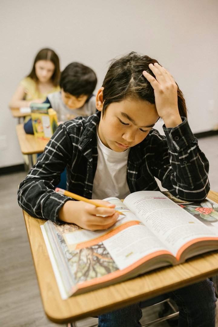 A pupil looks down at a textbook with a hand on their head, reflecting stress and emotional strain during school hours. | Source: Pexels