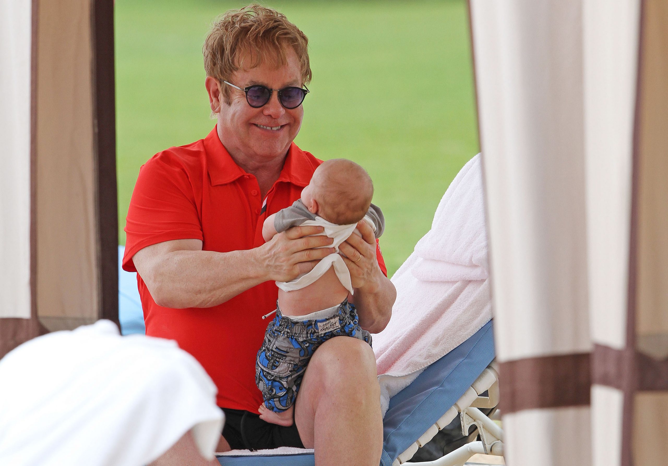 Sir Elton John shared a playful moment with baby Zachary John Furnish-John while on holiday in Honolulu, Hawaii, on February 21, 2011. Dressed casually in a bright red polo shirt and dark shorts, the singer lifted his infant son gently in his hands, smiling down at him with clear delight against a tropical backdrop and cabana setting.
