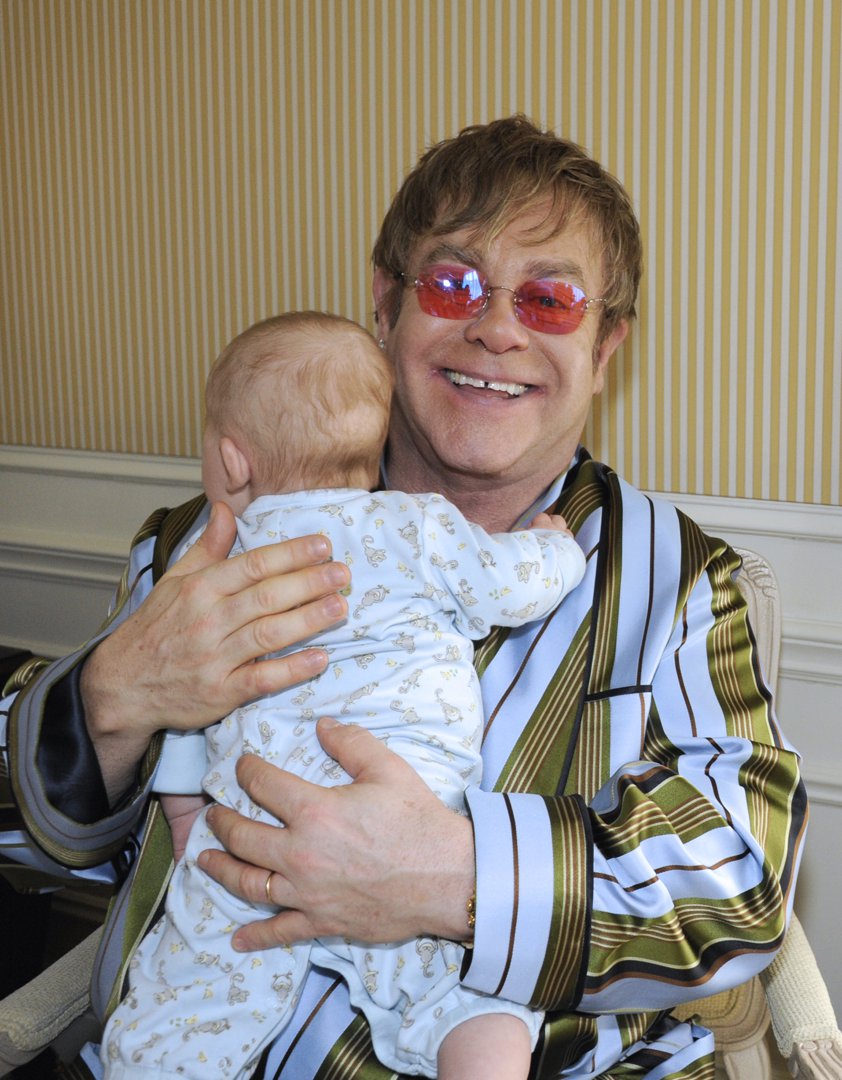 Sir Elton John cradled a baby Zachary John Furnish-John in his arms on April 18, 2011, smiling broadly as the infant rested against his chest. Dressed in a striped satin robe and his signature tinted glasses, the singer beamed with unmistakable pride, offering an early glimpse of the hands-on father he would later describe as his greatest role.