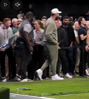 Travis Kelce, wearing a white cap, olive-green tracksuit, and white sneakers, walks along the edge of a turf field as security and event staff move nearby while a crowd watches from behind a barrier. | Source: Facebook/PageSix