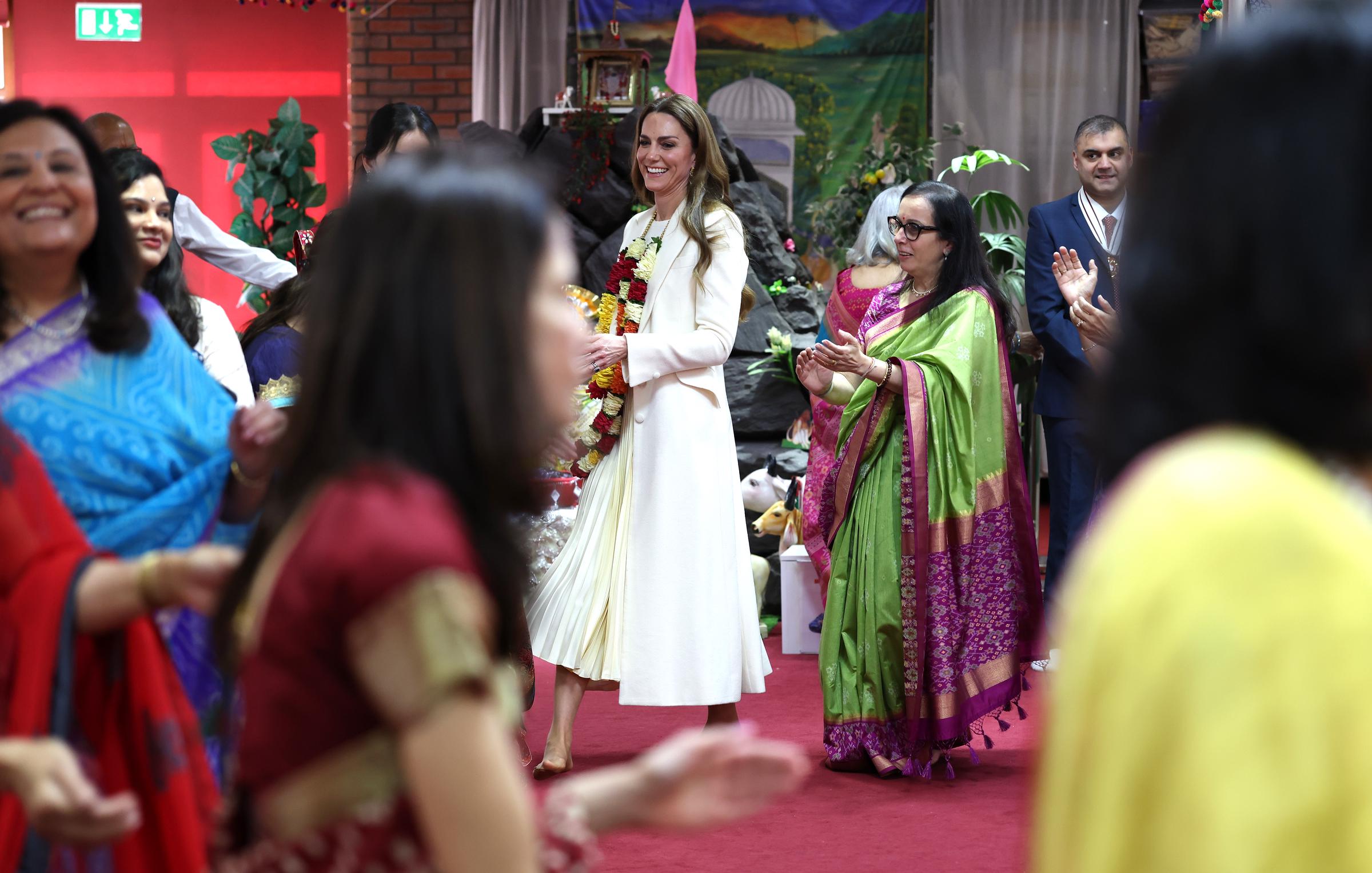 Catherine, Princess of Wales, smiles warmly as she is welcomed inside the Shreeji Dham Haveli Hindu Temple in Leicester, England, on March 5, 2026. Surrounded by colourfully dressed guests and temple members, the Princess of Wales walks through the hall wearing a vibrant flower garland, while those gathered applaud and greet her arrival.