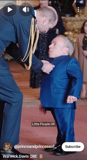 William, Prince of Wales, leans forward to carefully pin the OBE medal to Warwick Davis' royal blue jacket, the ornate surroundings of Windsor Castle forming a fitting backdrop for the occasion in a post shared on 11 March 2026. Warwick, who was born with Spondyloepiphyseal dysplasia congenita, a rare form of dwarfism, received the honour for his outstanding contributions to drama and charity. | Source: YouTube/The Prince and Princess of Wales