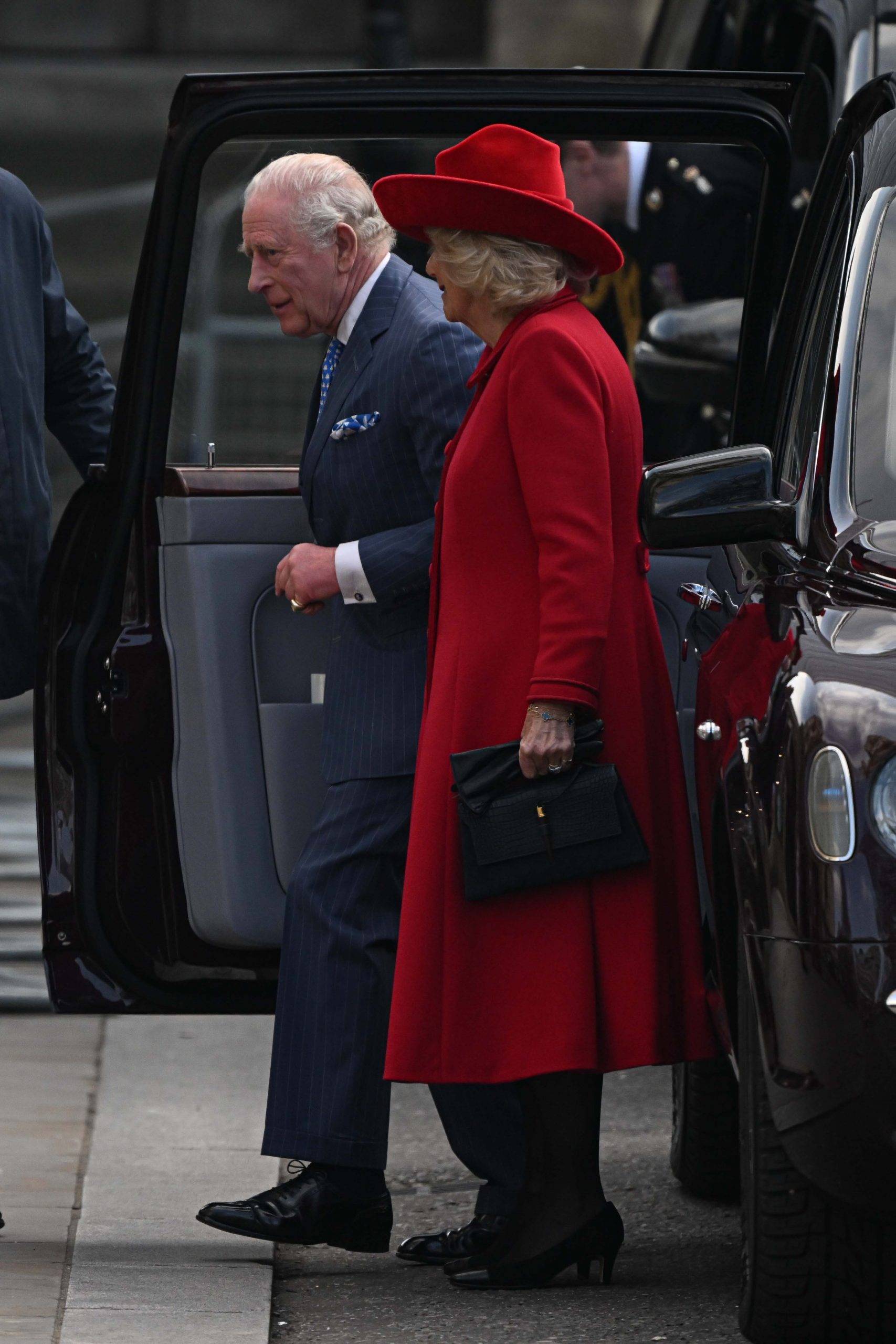 King Charles III, dressed in a sharp navy pinstripe suit with a blue patterned tie and pocket square, stepped from the royal car alongside Queen Camilla, who turned heads in a vivid red coat, matching wide-brimmed hat, and black leather handbag, as the pair arrived at Westminster Abbey for the Commonwealth Day service on 9 March 2026 in London &mdash; the last senior royals to enter the Abbey that afternoon.