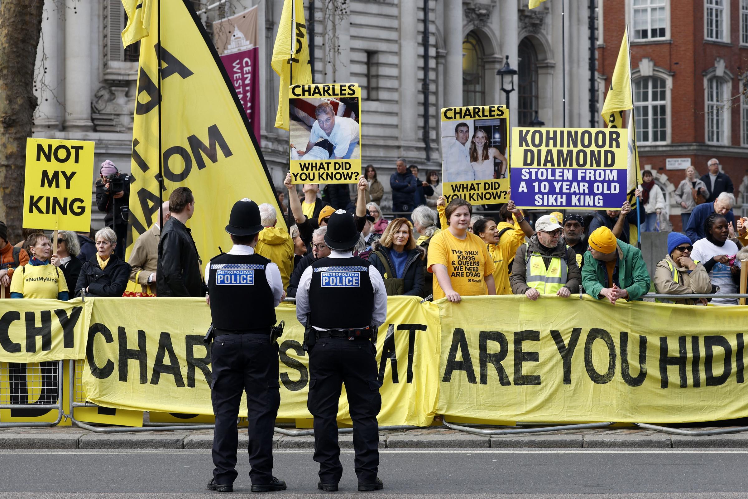 Two Metropolitan Police officers stood with their backs to the camera facing a crowd of anti-monarchist protestors behind bright yellow barriers outside Westminster Abbey on 9 March 2026, the demonstrators brandishing 