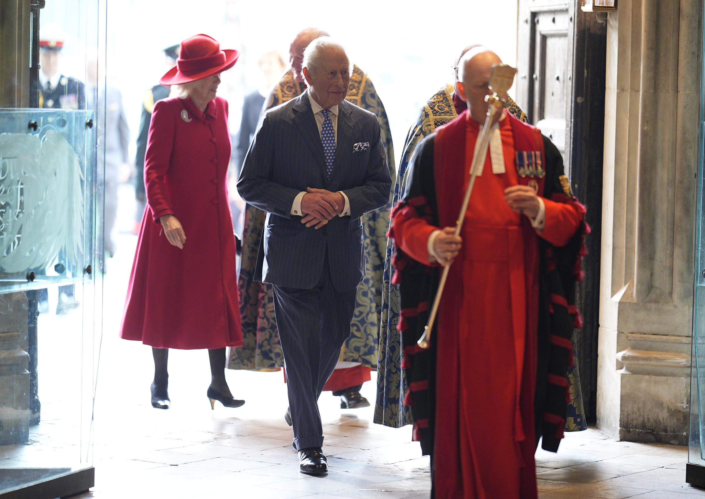 King Charles III, hands clasped calmly at his waist, walked through the entrance of Westminster Abbey led by a clergy member in ceremonial red robes and gold vestments, with Queen Camilla's vivid red coat a striking flash of colour as the pair processed into the church for the Commonwealth Day service on 9 March 2026.