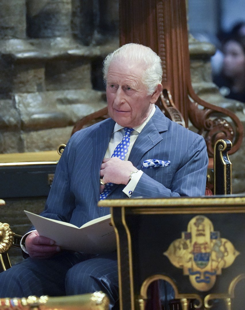 King Charles III sat in an ornate gold-trimmed ceremonial chair inside Westminster Abbey on 9 March 2026, his order of service booklet open in one hand while the other rested thoughtfully at his chest, his gold wristwatch catching the light alongside the blue patterned pocket square in his jacket as he followed the Commonwealth Day service with quiet concentration.