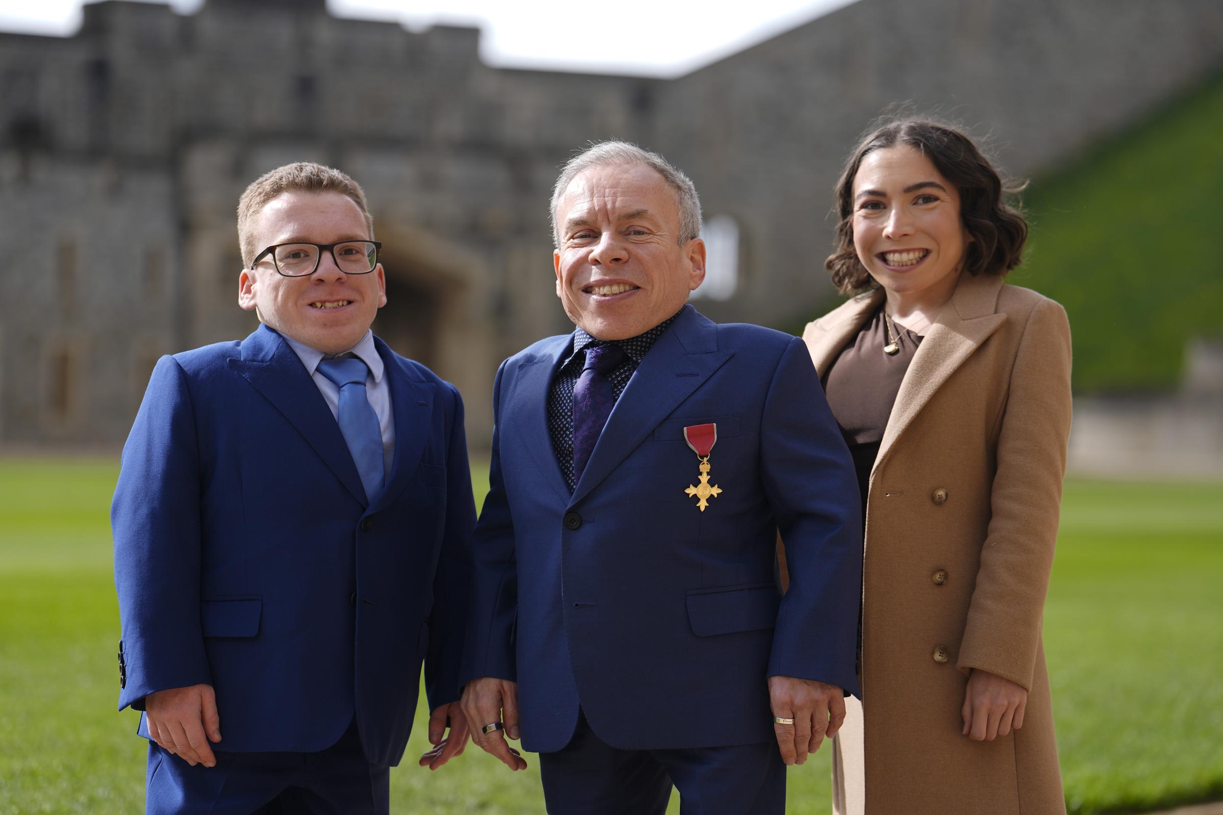 Warwick Davis is flanked by his son, Harrison Davis, 23, and daughter, Annabelle Davis, 28, on the grounds of Windsor Castle on 11 March 2026, all three grinning broadly for the camera with the castle's ancient stone walls stretching out behind them. It is a family portrait with real weight to it &mdash; the children who stood by their father through grief were also there to witness one of the proudest moments of his career.