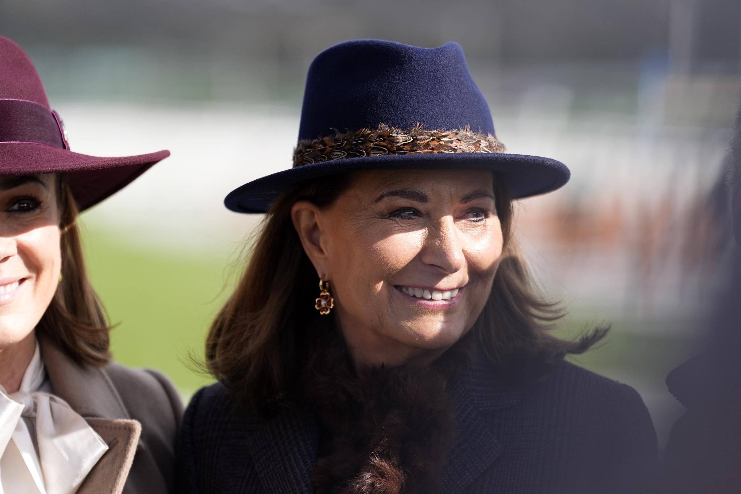 Beaming in the Gloucestershire sunshine, Carole Middleton sports the feather-trimmed Hicks & Brown blue fedora &mdash; the very hat last seen on the head of the Princess of Wales at Sandringham in 2020 &mdash; its distinctive pheasant-feather band catching the light as her floral drop earrings frame a radiant, rosy-lipped smile at Cheltenham Racecourse on 11 March 2026.
