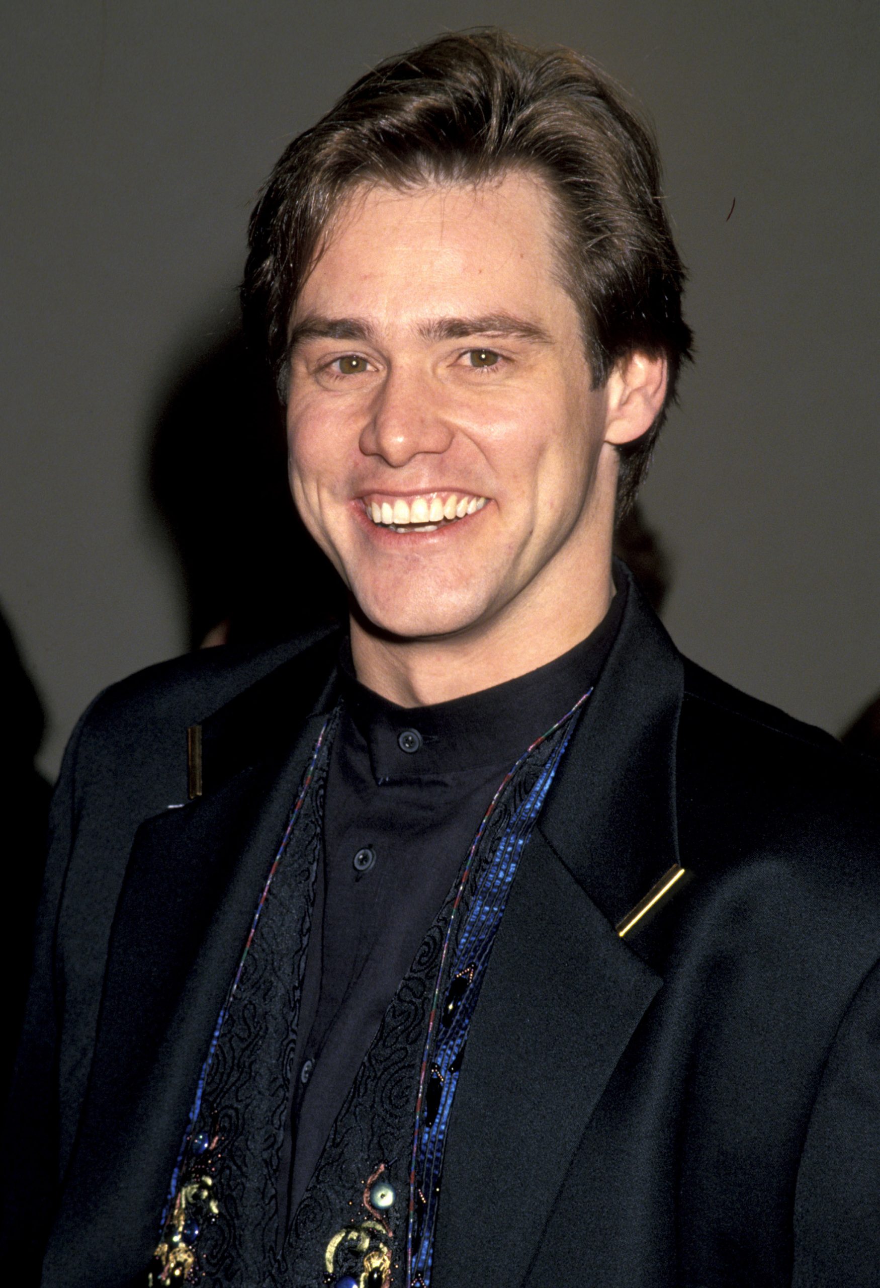 Beaming with a wide, boyish grin, Jim Carrey exudes early-career charm in a sleek black tuxedo layered over an intricately patterned vest at the &ldquo;Ace Ventura: Pet Detective&rdquo; premiere. His neatly styled hair and playful confidence reflect a rising star stepping into the spotlight with unmistakable presence.
