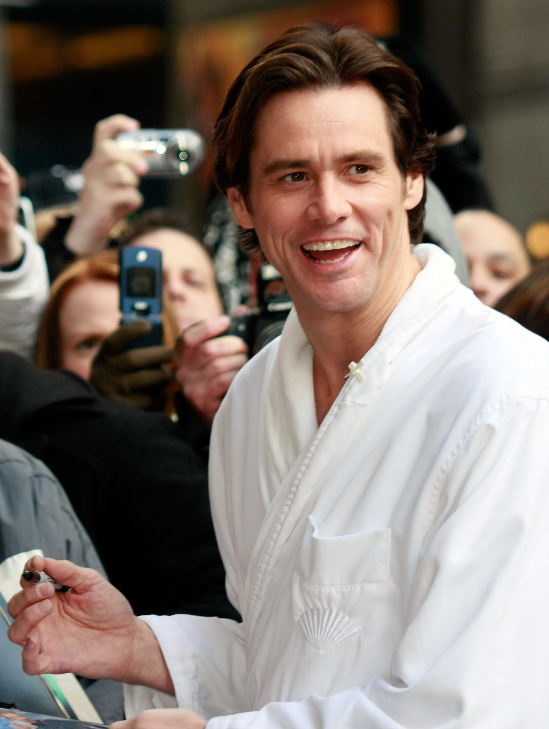 Wearing a crisp white robe-style top, Jim Carrey beams mid-laugh as he signs autographs outside the Ed Sullivan Theater. Fans crowd in behind him, holding up flip phones and cameras to capture the moment. He is pictured during a visit to the Late Show with David Letterman in New York City.