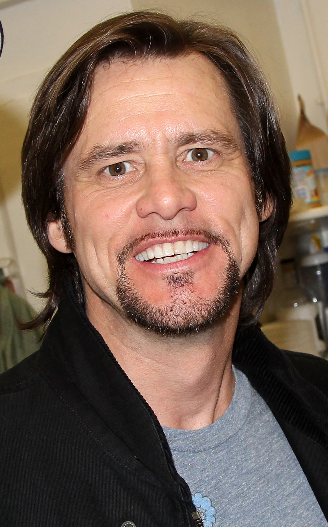 Sporting a neatly trimmed goatee and shoulder-length hair, Jim Carrey smiles warmly for a close-up backstage moment. He is dressed casually in a gray tee layered beneath a dark jacket. The actor poses at the hit musical 