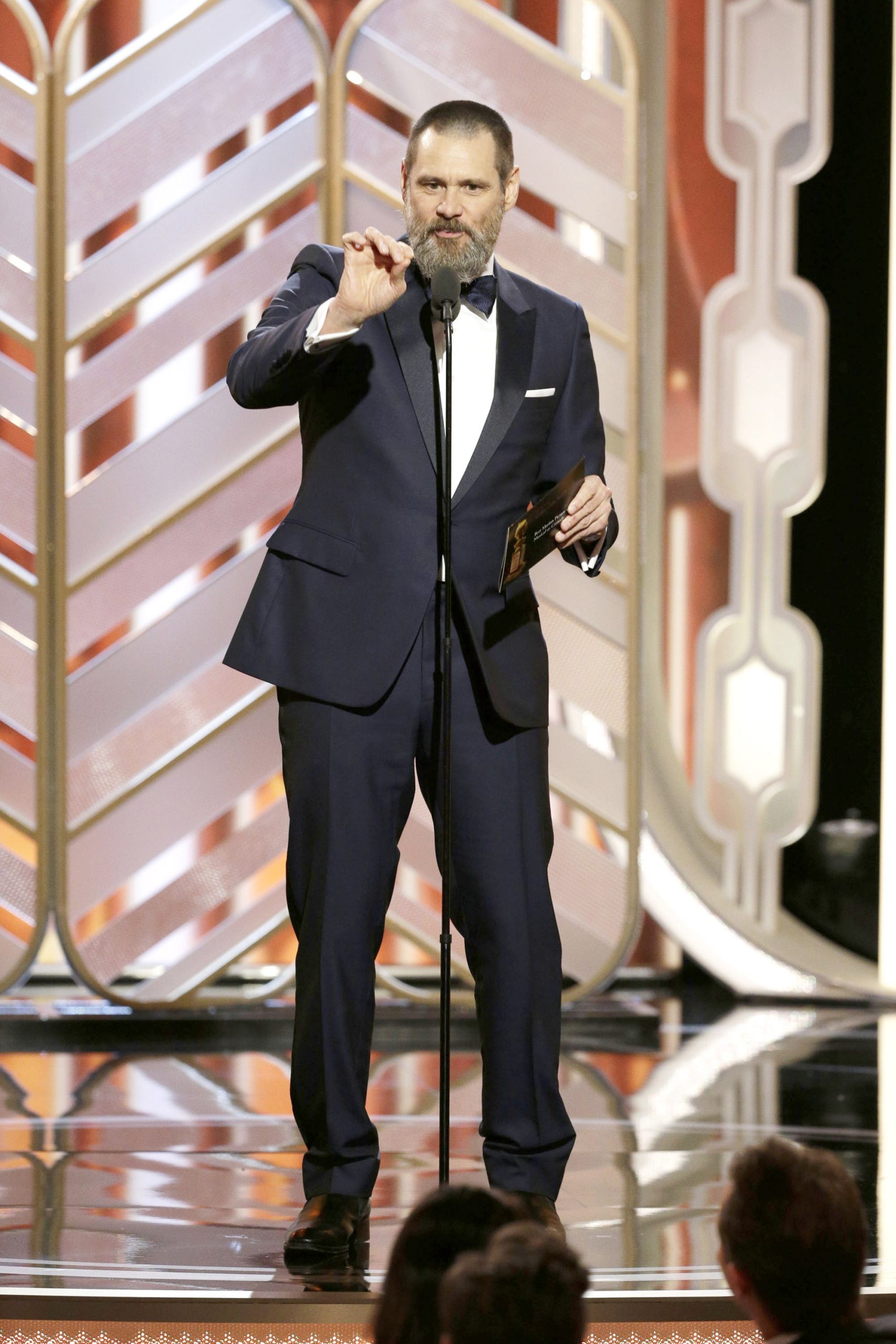 Dressed in a sharp navy tuxedo and bow tie, Jim Carrey commands the stage with a poised, mid-gesture delivery at the microphone. He holds an envelope in one hand while addressing the audience with his signature animated presence.