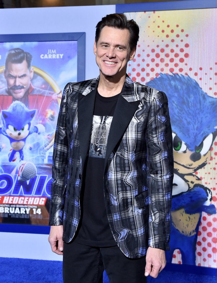 Wearing a patterned plaid blazer over a graphic black tee, Jim Carrey flashes a relaxed smile on the blue carpet. Behind him, vibrant posters of 