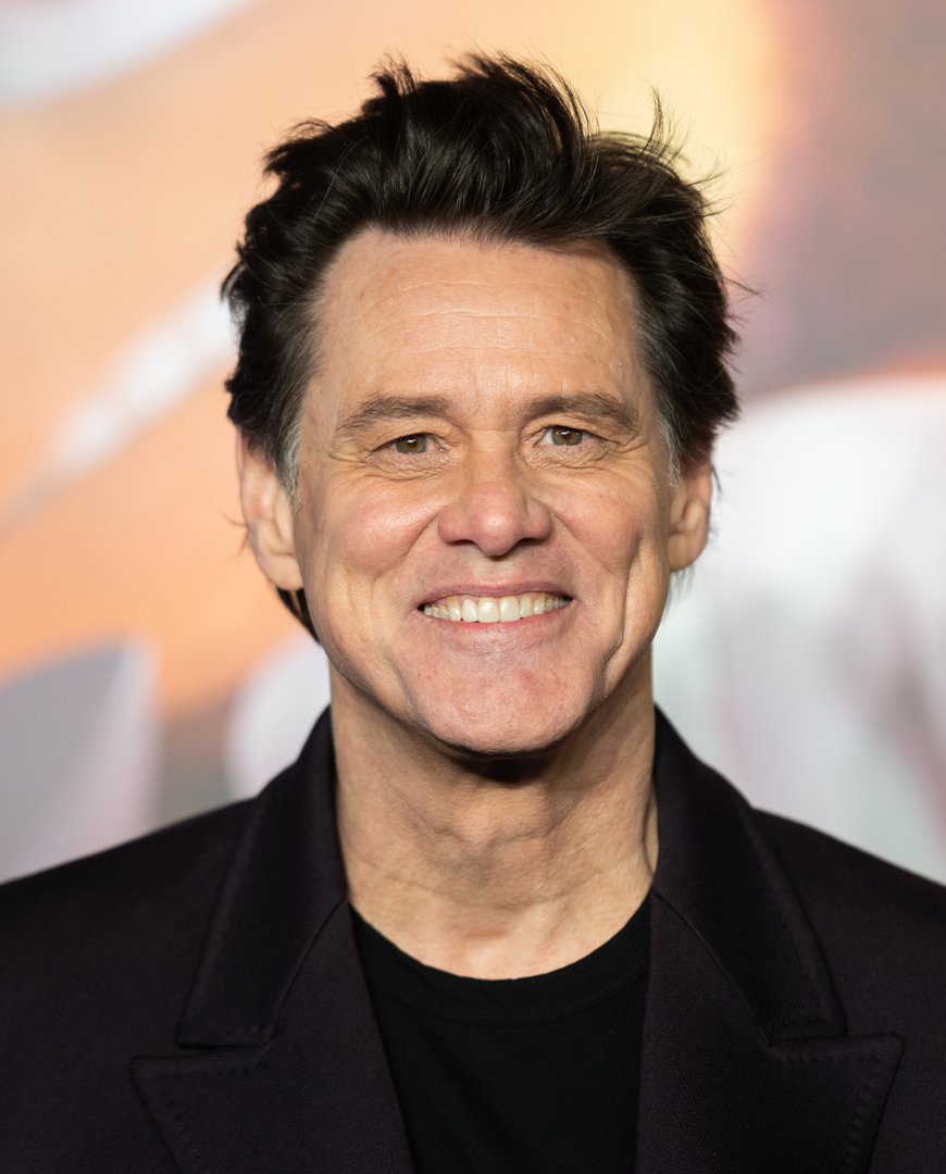 Smiling warmly for the cameras, Jim Carrey appears in a sleek dark ensemble with softly tousled hair. The blurred premiere backdrop casts a warm glow that frames his unmistakable grin. He attends the 