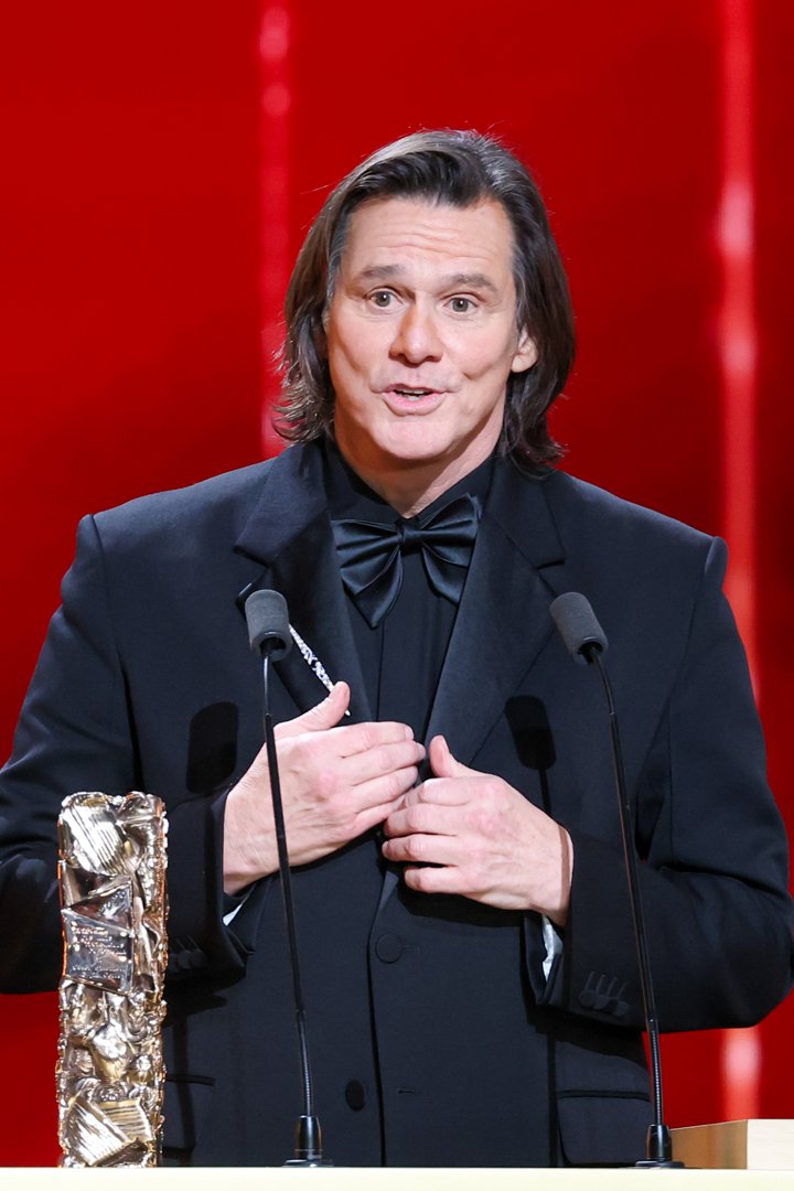 Dressed in a sharp black tuxedo, Jim Carrey stands at the podium with the Honorary C&eacute;sar Award beside him. He gestures mid-speech between twin microphones against a bold red stage backdrop. The actor accepts the honor during the 51st C&eacute;sar Film Awards.