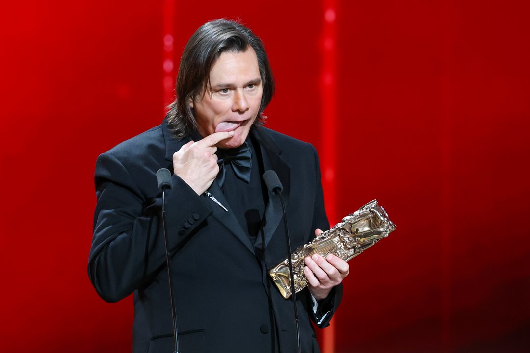 Holding his Honorary C&eacute;sar Award, Jim Carrey playfully points to his tongue mid-speech. Dressed in a classic black tuxedo, he stands between two microphones against a striking red backdrop. The light hearted moment unfolds onstage at the 51st C&eacute;sar Film Awards at L'Olympia in Paris, France.