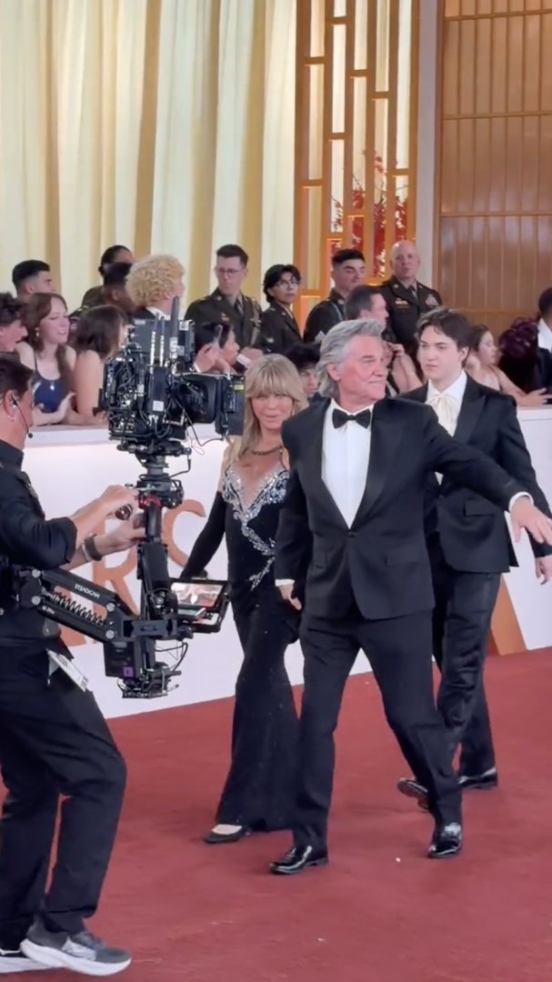 Kurt Russell gestures toward someone in the crowd as Goldie Hawn walks behind him, smiling while making her way through the packed arrivals area. | Source: Instagram/enews