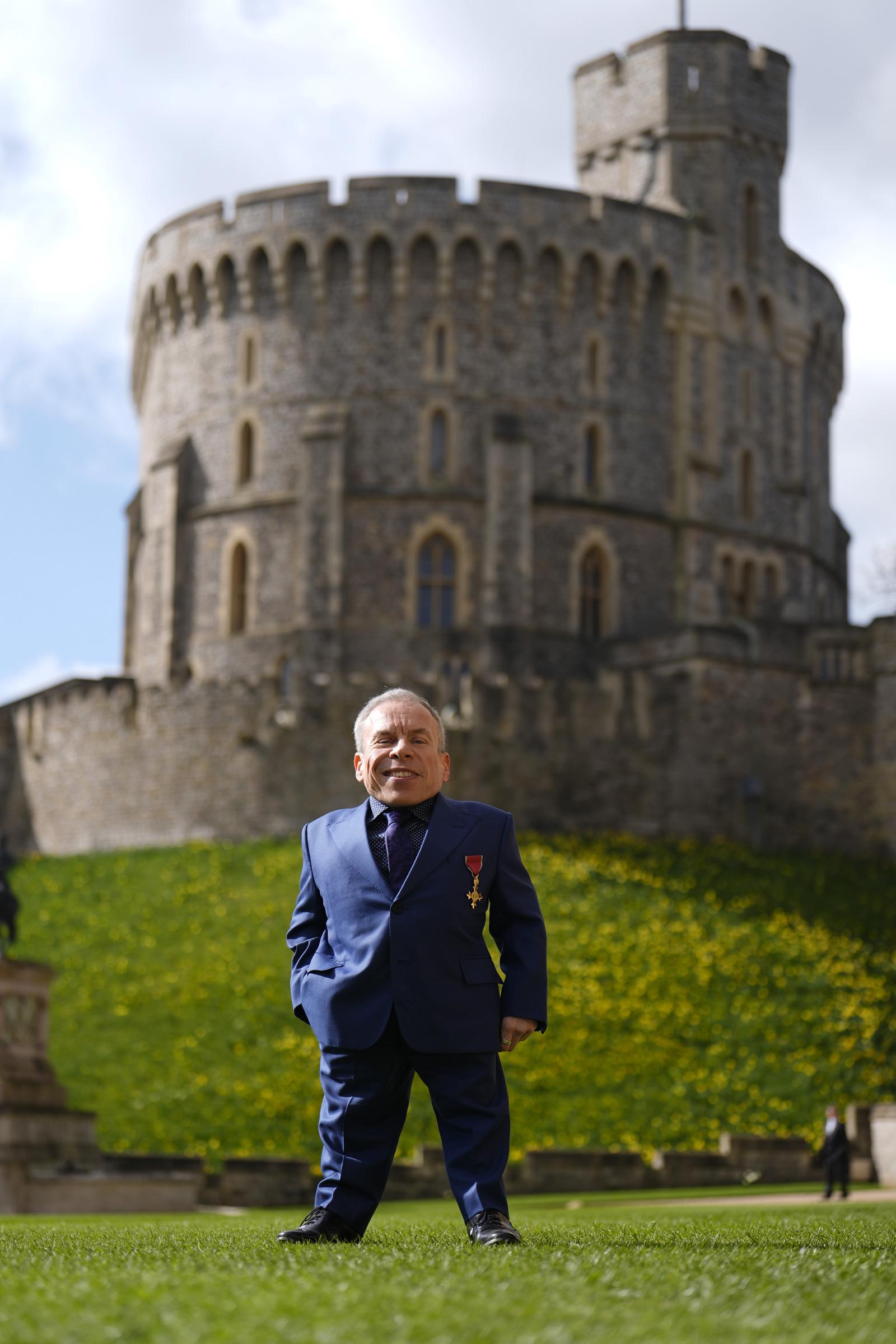 A beaming Warwick Davis poses on the grounds of Windsor Castle following his investiture on 11 March 2026, his OBE medal catching the light against his royal blue suit as the castle's iconic Round Tower rises behind him. The broad smile on his face says everything — this was a day four decades in the making, honouring a career that has taken him from a galaxy far, far away to the corridors of Hogwarts and beyond.