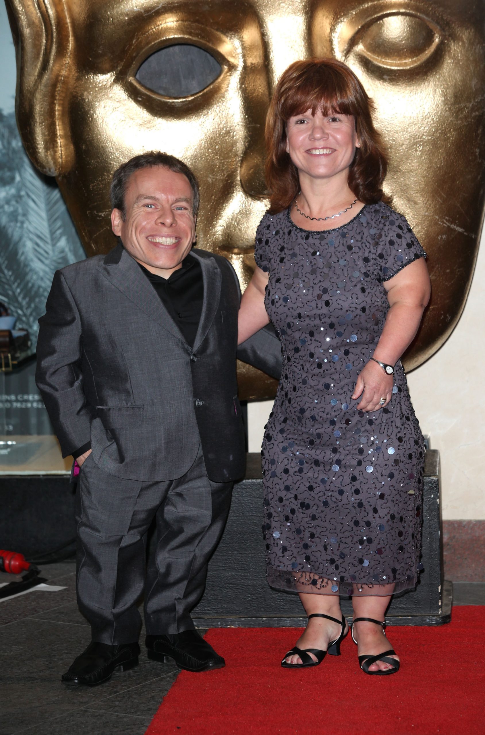 Warwick and Samantha Davis link arms on the red carpet at the British Academy Children's Awards in London, England, on 27 November 2011, both smiling broadly in front of the iconic golden BAFTA mask — a fitting backdrop for a couple who were no strangers to the industry's most prestigious stages. When Warwick received the BAFTA Fellowship years later, it was Samantha he chose to dedicate it to, describing it as