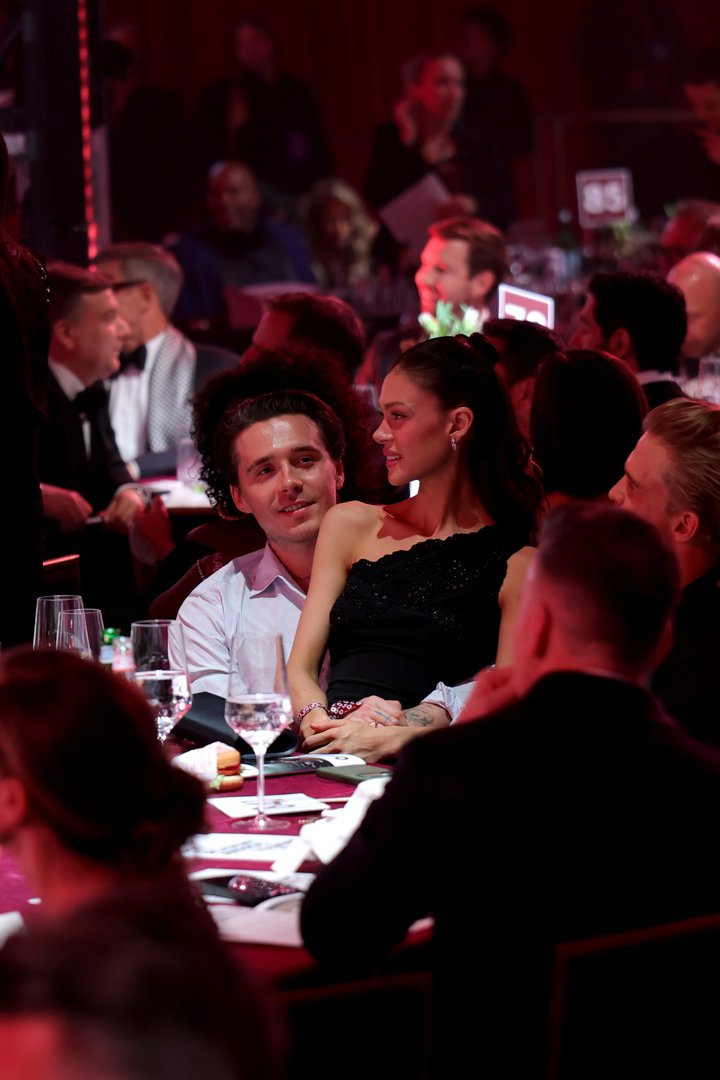 Across the table, in a more candid moment between the two, Nicola Peltz Beckham sits in Brooklyn Peltz Beckham's lap as he it snapped turning, the pair leaning close with their hands clasped together on the burgundy tablecloth. Her hair is pulled back into a half up, half down hairstyle for this shot, and the warm red glow of the room fills the background, where other guests can be seen gathered around nearby tables in the packed venue.