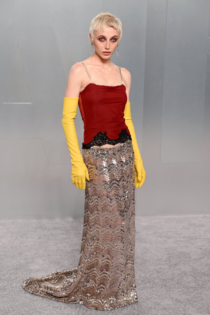 Emma Chamberlain wears a deep red lace-trimmed spaghetti-strap camisole top with black lace detailing at the hem, paired with a floor-length silver sequined chevron-patterned skirt — and bright yellow elbow-length rubber gloves — accessorised with long drop crystal earrings and a cropped platinum pixie cut.