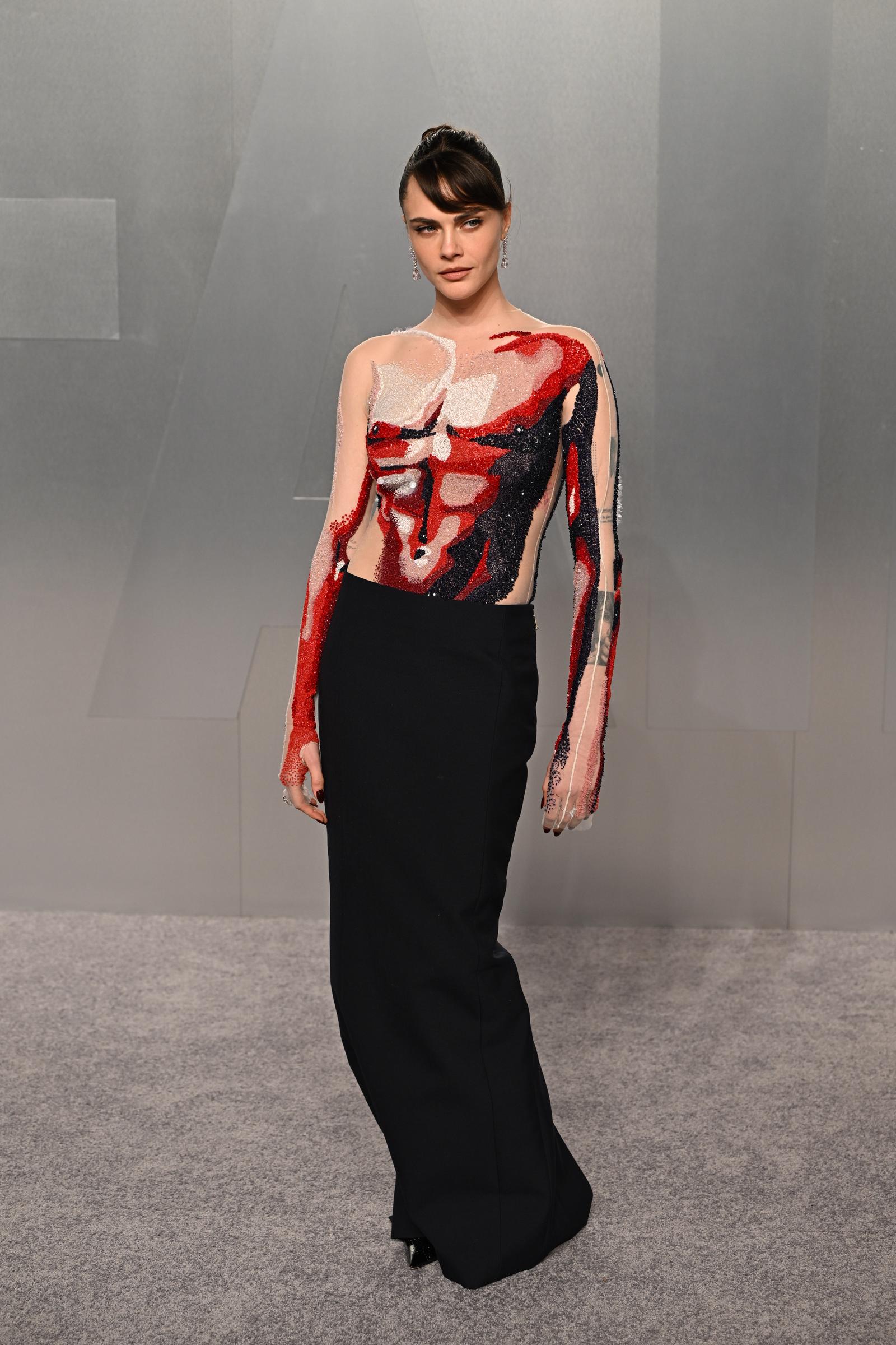 Cara Delevingne poses at the Vanity Fair Oscar Party in a sheer nude long-sleeved bodysuit decorated with abstract painterly motifs in red, white and black beading and sequins across the torso and arms, paired with a simple floor-length black column skirt and long diamond drop earrings.