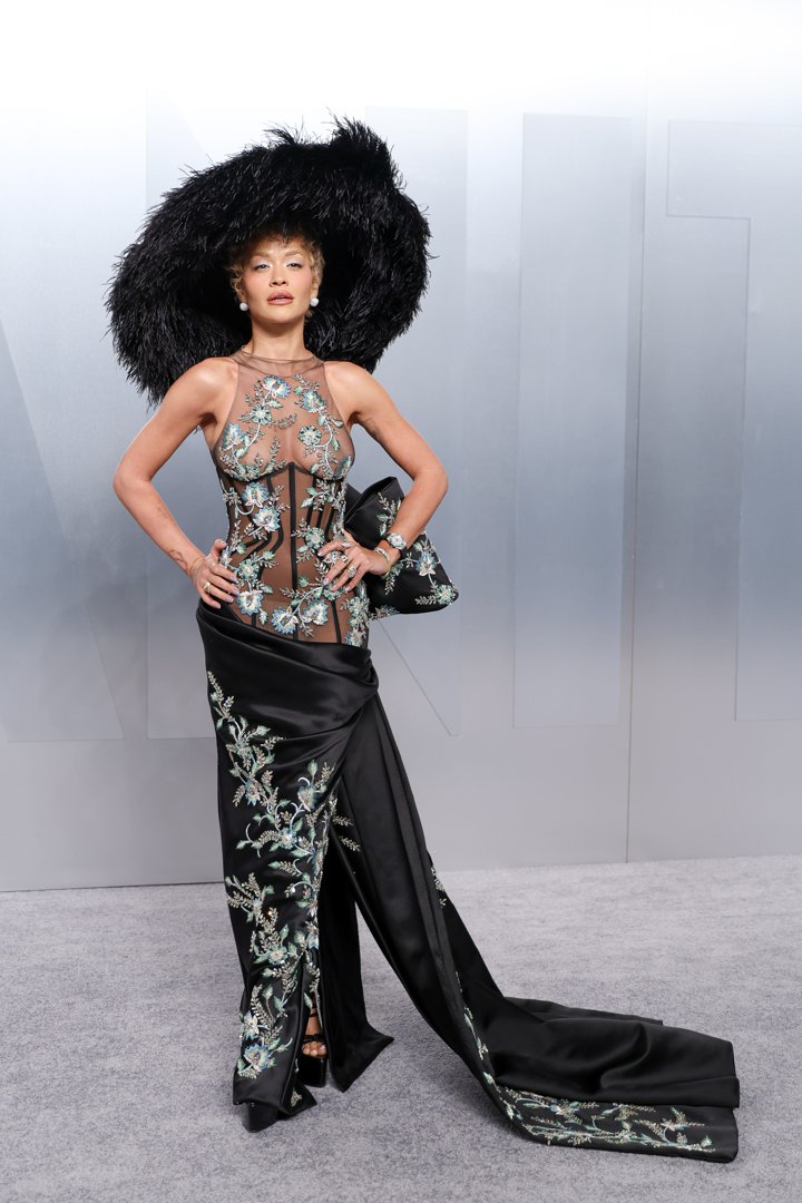 Rita Ora stands with hands on hips in a sheer nude halterneck corset bodice embroidered with iridescent floral motifs, a draped black satin skirt with matching floral embroidery and a sweeping train, paired with an oversized black feathered wide-brim hat, a black bow clutch and pearl stud earrings.