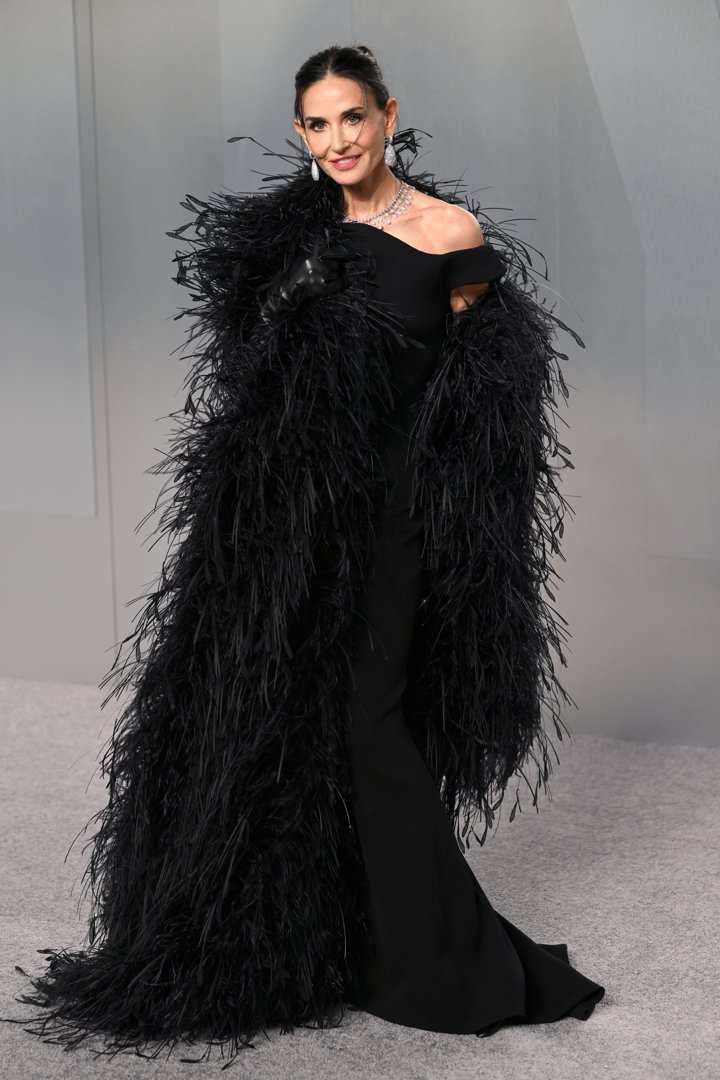 Demi Moore smiles for the cameras in a sleek black off-the-shoulder column gown almost entirely obscured by an enormous floor-length black feathered cape with voluminous feathering from shoulder to hem. She accessorised with a layered diamond necklace and matching teardrop earrings.