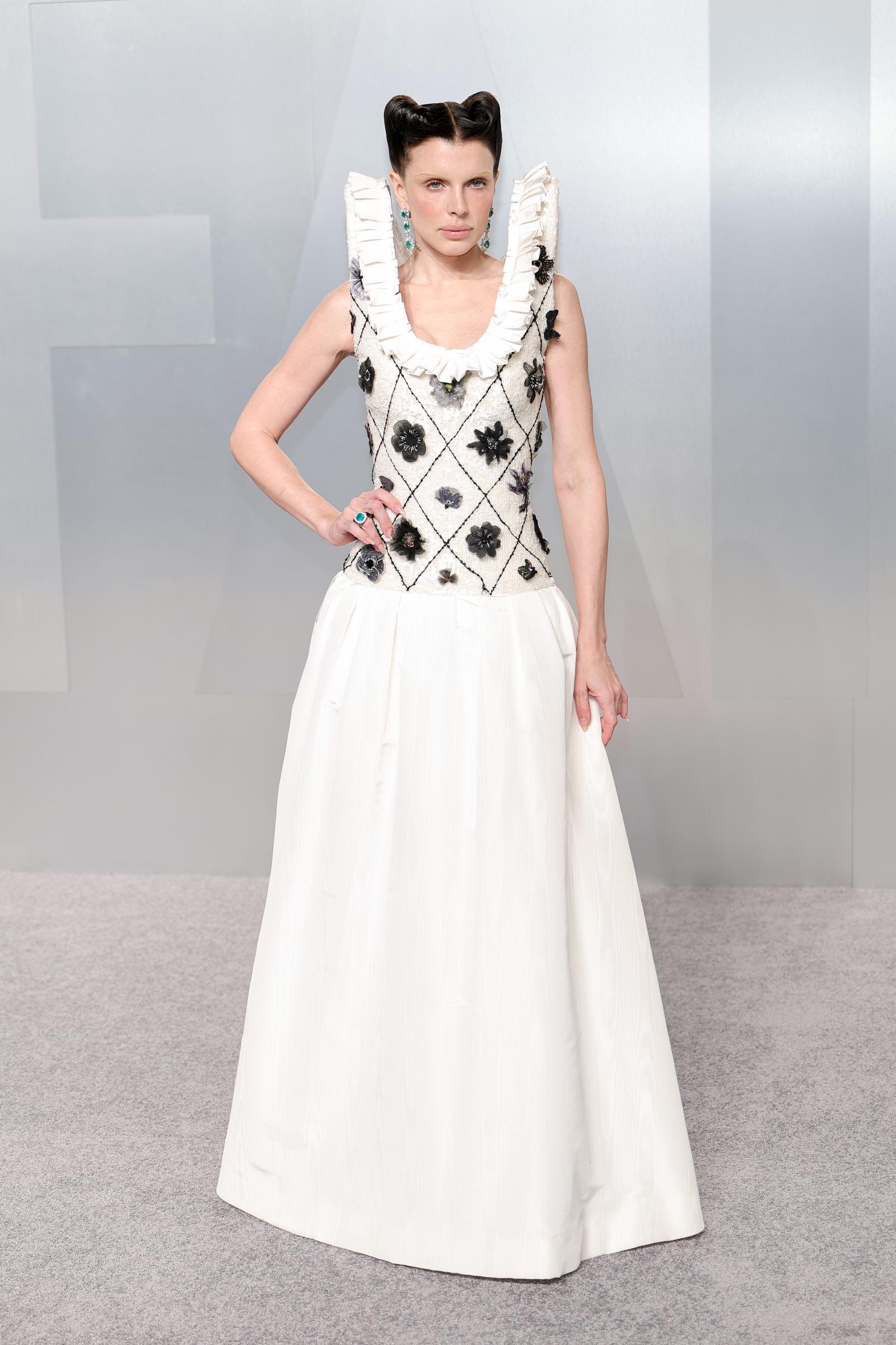 Julia Fox commands attention in a white drop-waist gown with a textured diamond lattice bodice embellished with 3D black floral appliqués, wide white ruffled shoulder straps, a full structured white A-line skirt, teal gemstone drop earrings and a matching cocktail ring. Her hair is styled in a dramatic sculptural pin-up updo.